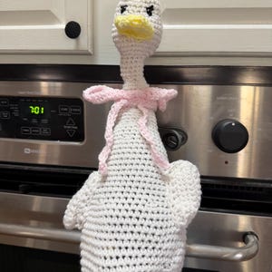 May include: A white crocheted goose toy with a yellow beak and feet, featuring black embroidered eyes and a light pink crocheted ribbon tied around its neck. The goose has a long neck and a rounded body, crafted with visible yarn stitches.