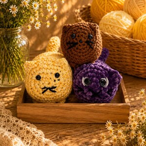 May include: A wooden tray holds three crocheted cat toys in yellow, brown, and purple. Each cat has black facial features and button eyes. Yellow yarn balls are in a basket behind the toys, and a vase of daisies is to the left.