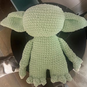 May include: A light green plush toy crafted from thick, textured yarn. The figure features a large, rounded head with prominent, wide-set pointed ears, a stout torso, long arms, and short legs with textured feet. The entire surface has a consistent knit.