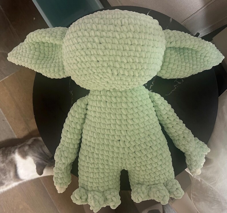 May include: A light green plush toy crafted from thick, textured yarn. The figure features a large, rounded head with prominent, wide-set pointed ears, a stout torso, long arms, and short legs with textured feet. The entire surface has a consistent knit.