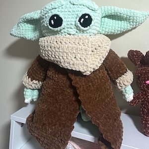 May include: A plush crochet doll featuring a light green head with large black eyes and pointed ears, wearing a textured beige scarf around the neck and a long brown coat with beige cuffs. The doll is crafted from soft, thick yarn material.
