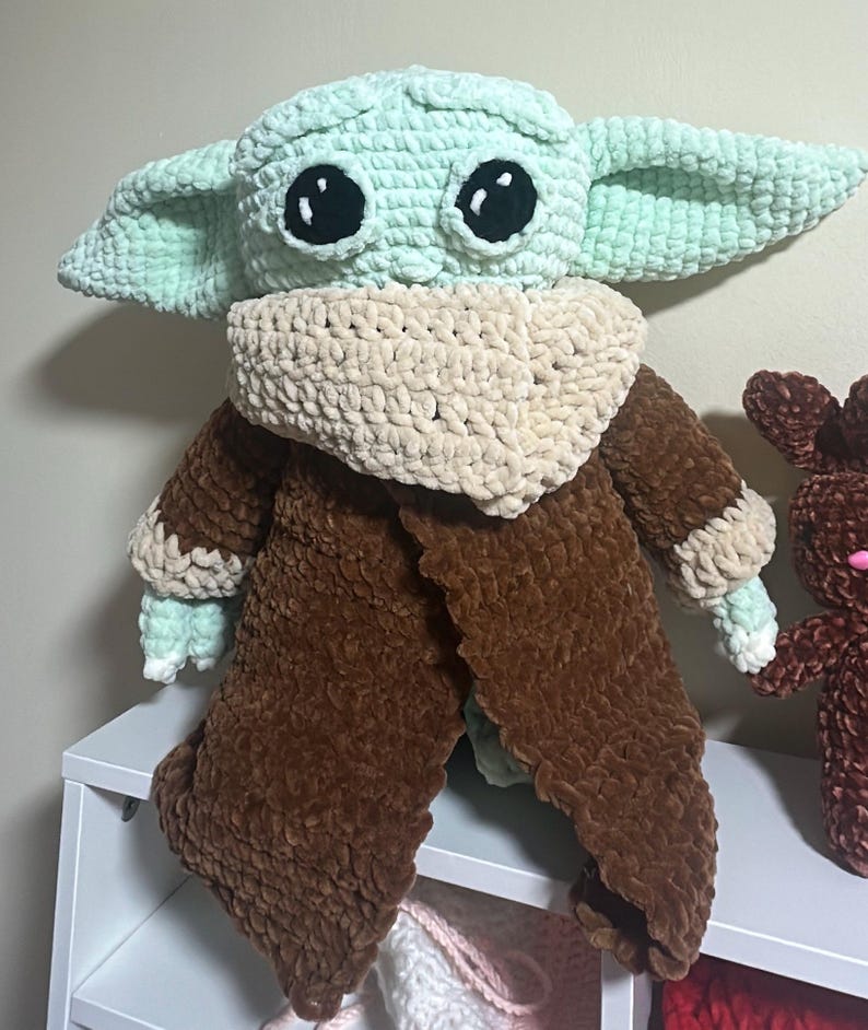 May include: A plush crochet doll featuring a light green head with large black eyes and pointed ears, wearing a textured beige scarf around the neck and a long brown coat with beige cuffs. The doll is crafted from soft, thick yarn material.