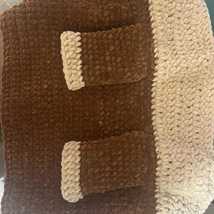 May include: A rectangular crochet bag made from thick, plush yarn in dark brown and cream colors. The bag features a solid dark brown body with two rectangular pockets on the front, each accented with a cream-colored trim, and a cream-colored side panel.
