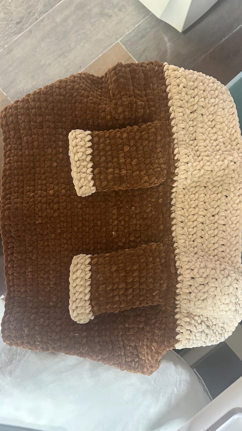 May include: A rectangular crochet bag made from thick, plush yarn in dark brown and cream colors. The bag features a solid dark brown body with two rectangular pockets on the front, each accented with a cream-colored trim, and a cream-colored side panel.