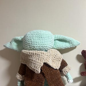 May include: A plush crochet doll featuring a light green head with large pointed ears, wearing a dark brown robe with a thick, textured beige collar. The doll is crafted from soft, chunky yarn and is positioned facing away from the viewer.