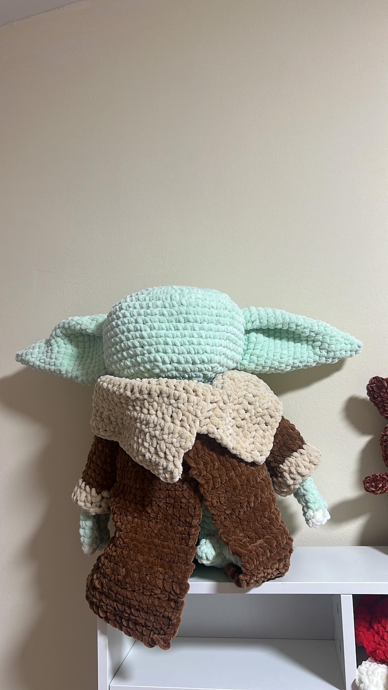May include: A plush crochet doll featuring a light green head with large pointed ears, wearing a dark brown robe with a thick, textured beige collar. The doll is crafted from soft, chunky yarn and is positioned facing away from the viewer.