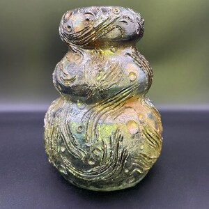May include: A small, three-tiered gourd-shaped vase with a metallic, iridescent finish. The surface features raised, swirling, wave-like textures and small circular indentations throughout, blending shades of dark green, gold, and bronze.