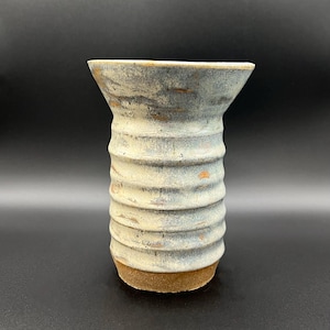 May include: A handmade ceramic vase with a textured, ribbed design. The vase features a light blue and cream color scheme with brown accents. The base is a darker brown. The vase has a flared top and is suitable for displaying flowers or as a decorative piece.