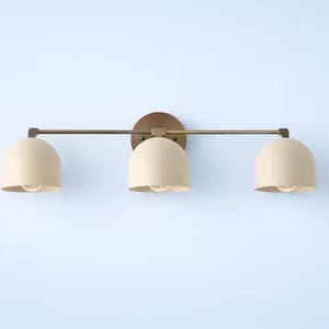 May include: A three-light vanity light with a bronze finish. It features three cream-coloured, dome-shaped shades. The light fitting is mounted on a light blue wall.