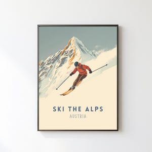 Könnte beinhalten: Gerahmtes Poster im Vintage-Stil mit einem Skifahrer, der einen schneebedeckten Berghang hinunterfährt. Der Skifahrer trägt eine rote Jacke und einen schwarzen Helm. Das Poster zeigt "SKI THE ALPS AUSTRIA" in blauer Schrift auf cremefarbenem Hintergrund.