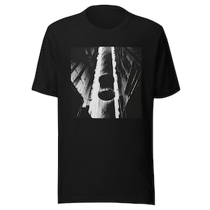 May include: A black short-sleeve t-shirt featuring a square graphic print on the chest. The print displays a high-contrast black and white architectural scene of a deep, narrow vertical shaft with a cylindrical structure suspended in the center.