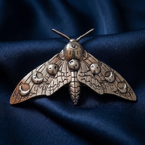 May include: A silver-toned metal moth brooch featuring an engraved eye on the thorax and various moon phase symbols across the wings. The moth has two antennae, a segmented body, and intricate line work detailing the wing patterns and textures.