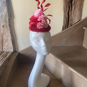 Pode incluir: Um fascinator vermelho com um arranjo floral e detalhes de fita. O adorno de cabeça apresenta uma base redonda, adornada com flores rosa e cachos de fita vermelha. O chapéu é exibido em uma cabeça de manequim branca.