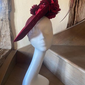 Pode incluir: Um fascinator em tom bordô profundo com uma aba larga e inclinada e um grupo de flores de tecido vermelho-escuro a condizer. A peça é adornada com um fino detalhe de arame curvo que se estende para cima e para fora do arranjo floral.