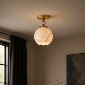 May include: A ceiling-mounted light fixture featuring a round, translucent, cream-colored stone globe shade with natural veining. The shade is suspended from a circular brass canopy by a short metal rod and a decorative ring connector.