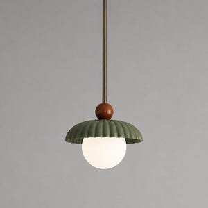 Op de afbeelding: Een hanglamp met een lange, dunne, bronskleurige metalen stang die uit een ronde plafondbevestiging komt. Het bevat een kleine, gladde, donkerbruine houten bol boven een brede, geribbelde, saliegroene kap en een witte glazen bol.