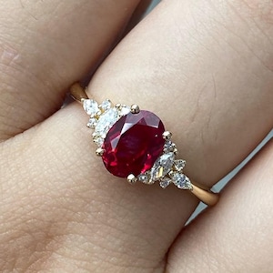 May include: A gold ring featuring a central oval-cut deep red ruby gemstone. The ruby is flanked on both sides by a cluster of small, clear, marquise-cut and round-cut accent stones set in a delicate gold band, creating a symmetrical design.