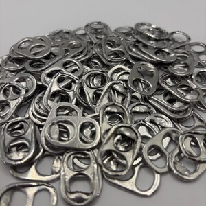 May include: A large pile of silver-colored aluminum soda can tabs. Each tab features a distinct oval-shaped ring and a smaller circular opening, with a metallic finish and smooth, rounded edges. The collection consists of numerous individual tabs.