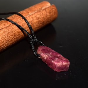 May include: A raw, deep pinkish-red gemstone pendant suspended from a thin black cord. The stone has an irregular, unpolished, and textured surface. The black cord is knotted at the top of the stone, creating a simple necklace design.