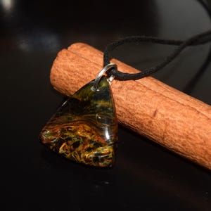 May include: A triangular pendant featuring a polished, multi-colored stone with swirling patterns of deep blue, gold, and black. The pendant is attached to a thin black cord and rests against a textured, cylindrical brown wooden element.