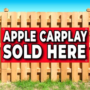 May include: A rectangular red banner with white text that reads APPLE CARPLAY SOLD HERE. The banner features four metal grommets in the corners and is mounted against a vertical wooden fence with a clear blue sky visible at the top edge.