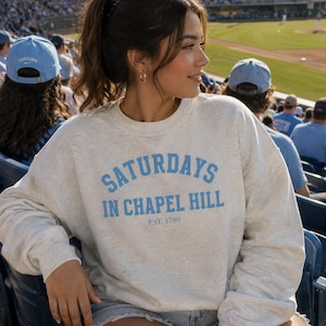 Pu&ograve; includere: Una felpa grigio chiaro con la scritta "SATURDAYS IN CHAPEL HILL" in azzurro, con "EST. 1789" sotto. La felpa &egrave; indossata da una persona a una partita di baseball. Le persone sullo sfondo indossano cappellini da baseball Carolina.