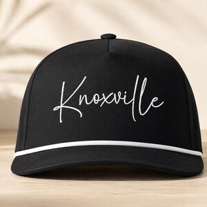Puede incluir: Una gorra de béisbol negra con la palabra Knoxville bordada en letra cursiva blanca en el panel frontal. La gorra incluye un cordón trenzado blanco que recorre horizontalmente la base de la corona, justo encima de la visera plana.