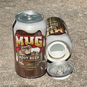 May include: A Mug Root Beer soda can with a silver pull-tab lid. The can features a brown background, a bulldog illustration, and the text MUG ROOT BEER NO CAFFEINE. A second can is lying on its side with the top removed, revealing a white inner liner.
