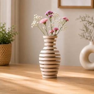 Puede incluir: Un jarrón de madera de color claro con un diseño de listones horizontales, que presenta un cuello estrecho y un cuerpo redondeado más ancho. El jarrón contiene un pequeño ramo de delicados claveles rosas y pequeñas flores blancas.