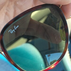 May include: Ray-Ban sunglasses with a tortoiseshell frame and green-tinted lenses. The brand name is visible on the lens. The sunglasses are held by a person.