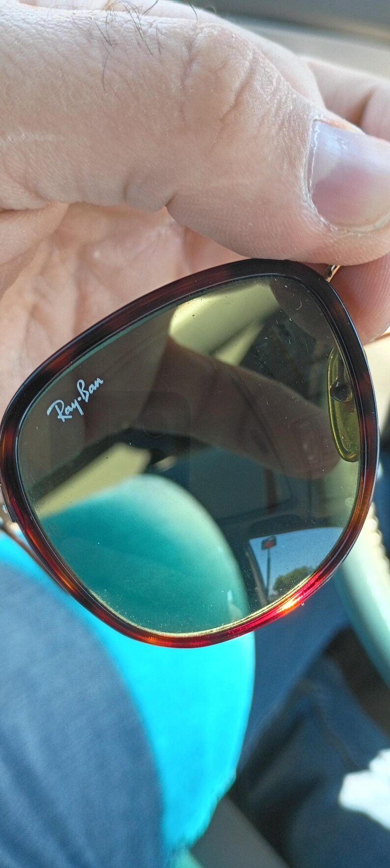 May include: Ray-Ban sunglasses with a tortoiseshell frame and green-tinted lenses. The brand name is visible on the lens. The sunglasses are held by a person.