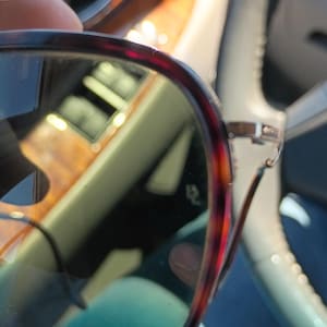 May include: Close-up of eyeglasses with a tortoiseshell pattern frame. The lenses are tinted, and the glasses are held in front of a blurred background, likely inside a vehicle. The frame has a reddish-brown and black pattern.