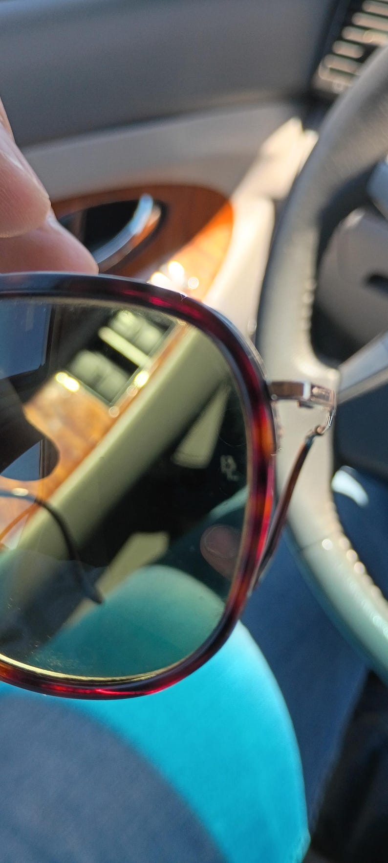 May include: Close-up of eyeglasses with a tortoiseshell pattern frame. The lenses are tinted, and the glasses are held in front of a blurred background, likely inside a vehicle. The frame has a reddish-brown and black pattern.