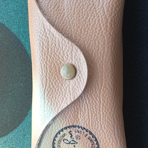 May include: Tan leather sunglasses case with a snap closure. The case has a textured surface and a circular logo with text that reads "Ray-Ban Sunglasses by Bausch & Lomb".