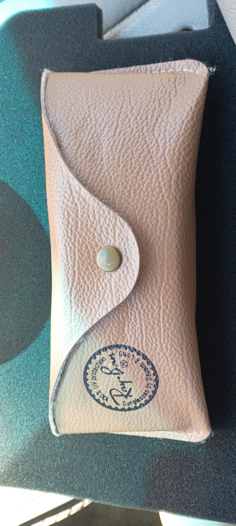 May include: Tan leather sunglasses case with a snap closure. The case has a textured surface and a circular logo with text that reads "Ray-Ban Sunglasses by Bausch & Lomb".