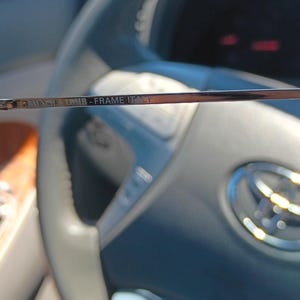 May include: Close-up of the frame of a pair of eyeglasses. The text "Bausch & Lomb - Frame Italy" is visible on the frame. The background is a car interior, including the steering wheel.