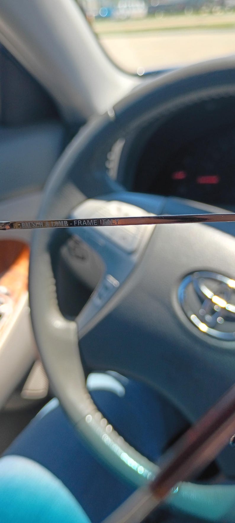 May include: Close-up of the frame of a pair of eyeglasses. The text "Bausch & Lomb - Frame Italy" is visible on the frame. The background is a car interior, including the steering wheel.