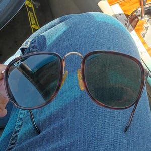 May include: Sunglasses with a brown tortoiseshell frame and dark lenses. The sunglasses are held against a background of blue denim jeans. The glasses have a classic, rounded shape and a metal bridge.