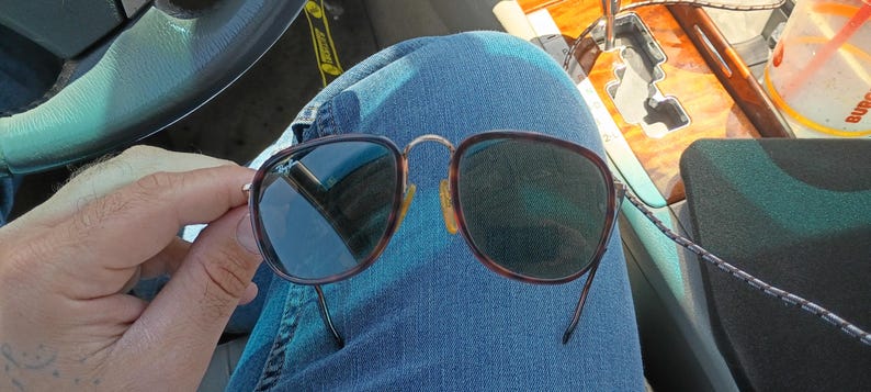 May include: Sunglasses with a brown tortoiseshell frame and dark lenses. The sunglasses are held against a background of blue denim jeans. The glasses have a classic, rounded shape and a metal bridge.