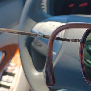 May include: Brown and gold-toned eyeglasses with a dark rim and green lenses. The glasses are held in front of a car's steering wheel, with the text "W0870" visible on the frame.