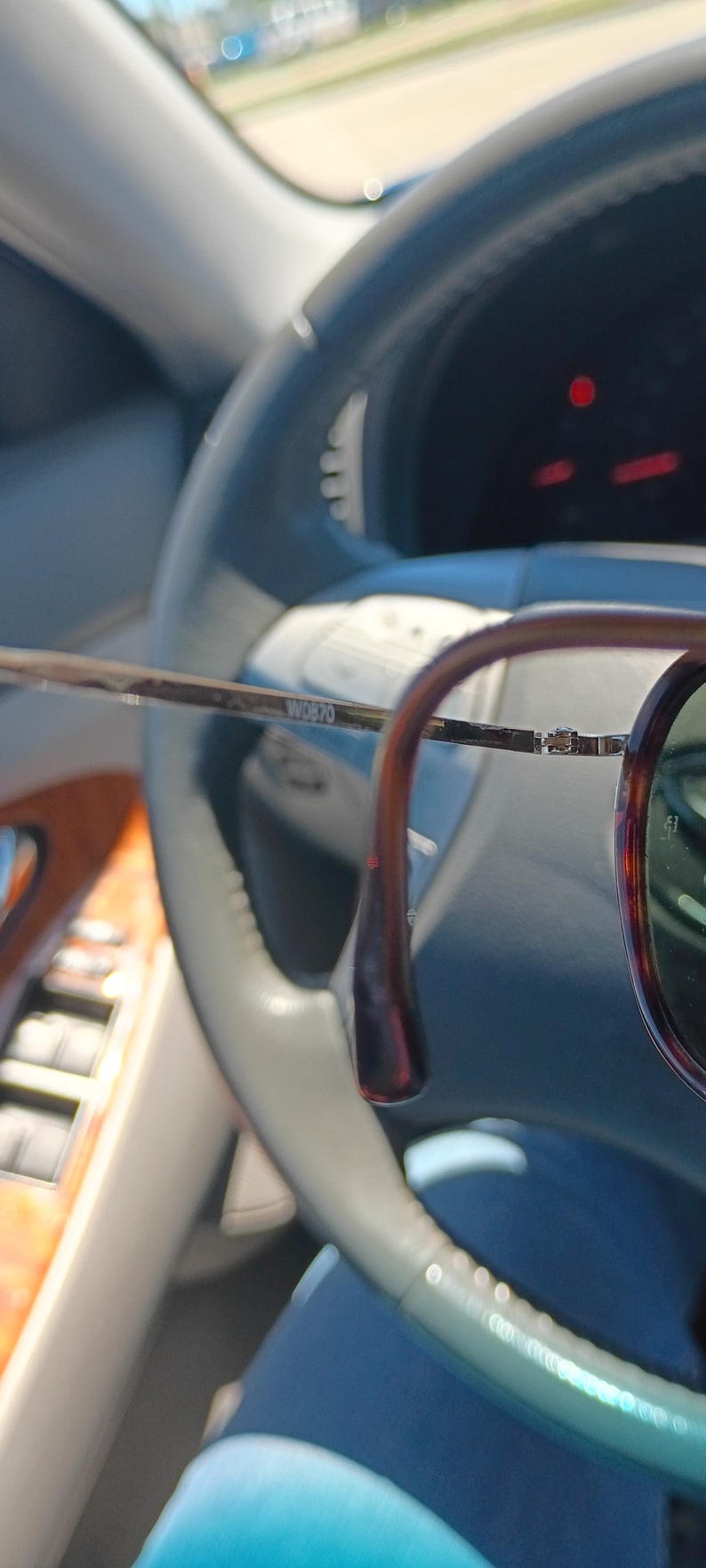 May include: Brown and gold-toned eyeglasses with a dark rim and green lenses. The glasses are held in front of a car's steering wheel, with the text "W0870" visible on the frame.