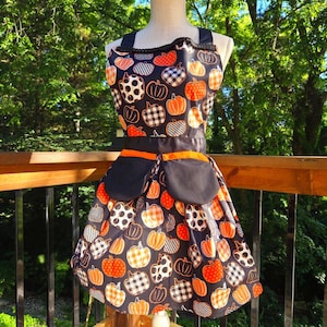 May include: A black apron featuring a repeating pattern of pumpkins in orange, white, and black with various textures like stripes and checks. The apron includes two solid black pockets with orange trim at the waist and a black ruffled neckline.