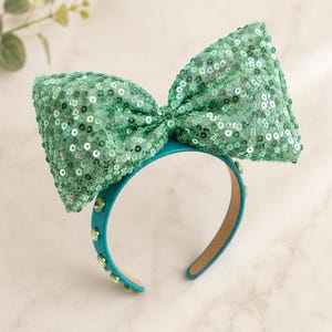 May include: A teal headband featuring a large, oversized bow covered in shimmering mint green sequins. The band itself is adorned with a series of small, round, light green faceted crystals embedded along the outer edge of the frame.