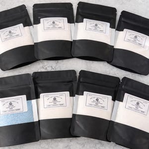 May include: Eight black stand-up pouches with clear windows, each containing a different product. The white labels feature the text The Tidy Laundry Co. and various scent names. One pouch contains light blue granules, while the others contain white.