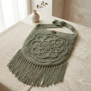 May include: A sage green crochet shoulder bag featuring a central circular mandala pattern, a single wide strap, a small brown wooden button closure at the top, and long fringe detailing along the bottom edge of the bag's main body.
