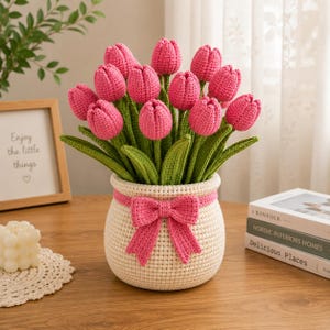 May include: A crochet bouquet of vibrant pink tulips with green stems and leaves arranged in a round, cream-colored crocheted pot. The pot is decorated with a matching pink crocheted bow wrapped around the upper section of the container.