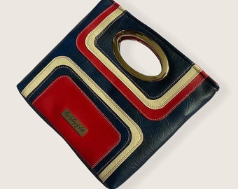 Puede incluir: Un bolso de mano azul, rojo y blanco con estampado geométrico y asa ovalada dorada. El bolso de mano tiene una etiqueta dorada con el texto "The Little Bag Lady".