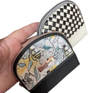 May include: Two small, handmade zippered coin purses. One features a black and white woven pattern, the other has a floral and bee design with the word "handmade" on a metal tag. The bottom of the floral purse is black.