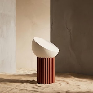 Può includere: Una scultura moderna con una parte superiore bianca a forma di mezza sfera appoggiata su una base cilindrica. La base è color terracotta con dettagli verticali a coste. La scultura è posta su sabbia, con pareti dai toni neutri sullo sfondo. Altezza: circa 30 cm.