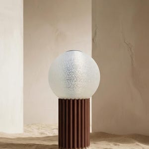 Può includere: Una moderna lampada da tavolo con paralume a globo in vetro smerigliato su una base cilindrica scanalata marrone scuro. La lampada è ambientata su uno sfondo neutro, creando un'estetica minimalista. Adatta per l'arredamento della casa.